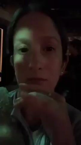 Here's a description of the video:

A woman with brown hair pulled back is seen in a dimly lit environment. She is wearing a light-colored shirt and a soft, textured jacket or sweater. The lighting is somewhat green-tinged. The woman is looking directly at the camera with a serious expression. Her hand is near her mouth, and she briefly moves it to show a ring on her finger. The background is dark, with indistinct shapes suggesting the presence of other people and perhaps an indoor setting, possibly a concert or performance venue due to the background noise of music.