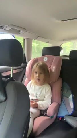 A young girl with blonde hair and a white shirt sits in a pink car seat in the back of a car. She is looking at the camera and appears to be talking. Trees and greenery are visible outside the car windows. The interior of the car is visible, including the back seats and headrests.