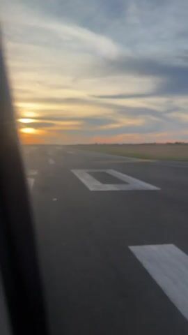 The video captures a view from an airplane window, looking out at a runway during what appears to be sunset. The runway is dark asphalt with white markings, including large rectangular shapes. The sun is low on the horizon, casting a warm glow and creating a soft light across the sky, which is filled with wispy clouds in shades of orange, yellow, and gray. Beyond the runway, a flat, open landscape with fields is visible. The interior of the airplane is partially visible, including a white seat cushion and the edge of the window frame. The video appears to be taken while the plane is taxiing on the runway, perhaps before takeoff or after landing.