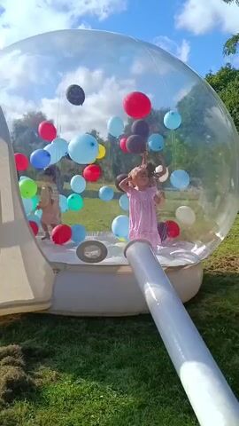 A young girl in a pink dress stands inside a large, clear inflatable bubble filled with balloons of various colors. She is looking towards the camera and has her hand raised to her forehead. Another person, partially visible, stands to her left within the bubble. A white tube extends from the side of the bubble, partially obscuring the girl's lower body. The bubble is situated outdoors on a grassy area, with trees and a blue sky visible in the background. Several balloons are floating in the air inside the bubble.