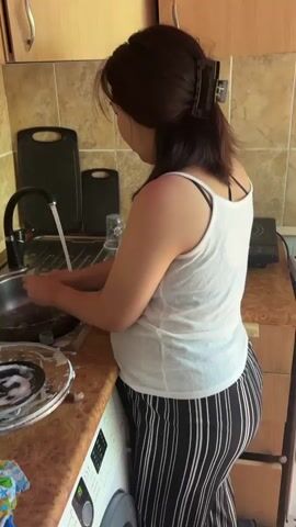 A woman with dark hair pulled back in a clip is standing at a kitchen sink, washing dishes. She is wearing a white tank top and black and white striped pants. The sink is stainless steel and has a black faucet. There are dishes in the sink, some with soap suds. A washing machine is visible to the left of the sink. The kitchen has tiled walls and wooden cabinets. A cutting board is leaning against the backsplash.
