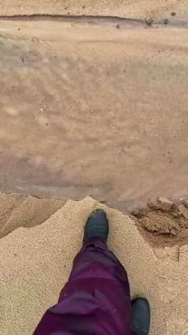 The video shows a person's legs, wearing purple pants and dark boots, standing on a sandy shore. The person is facing a small stream of water flowing through the sand, which leads to a larger body of water, possibly a lake or ocean, in the background. The stream has carved channels into the sand, and there is some debris, such as dried grass, in the water. The water is murky and appears to be flowing gently. The weather seems overcast. The camera is held at a high angle, looking down at the person's feet and the surrounding environment. The scene suggests a natural outdoor setting, possibly a beach or riverbank.