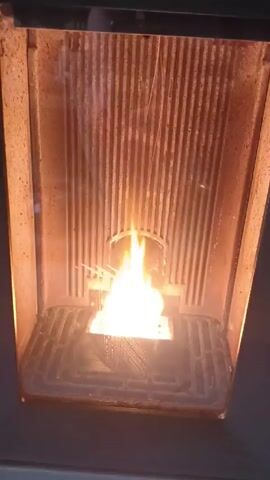 The video shows a pellet stove in operation. The fire burns brightly in the center of the grate, illuminating the interior of the stove, which is lined with vertical metal panels. There are a few sparks rising from the fire. The grate itself is a dark gray, while the interior of the stove is a tan or light brown color, possibly due to heat discoloration. The stove appears to be made of metal and is set against a dark background.