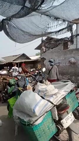 A bustling outdoor market scene unfolds. A motorcycle is parked in the foreground, laden with large sacks and green woven baskets, suggesting its use for transport. Several people are visible, some appearing to be vendors and others customers. One person in the background wears a white t-shirt with the number 79. Another individual is dressed in a red headscarf and dress. The market is covered with a black mesh netting to provide shade, and temporary structures made of wood and corrugated metal are present. The ground appears to be paved or dirt. The overall atmosphere is lively and suggests a typical day at a local marketplace.