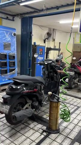The video shows the interior of a workshop, likely a motorcycle repair shop. In the foreground, a black scooter is parked on a stand, with a green hose and a collection drum for oil positioned next to it. The hose appears to be connected to the scooter, suggesting an oil change is in progress. In the background, a person wearing a dark shirt is visible, appearing to be working on a red scooter. There are two blue machines with digital displays in the background, and a large fan is mounted on the wall. The floor is tiled, and the walls are a light color. A large blue machine with compartments is on the left side of the frame. A folded blue metal door is visible in the middle of the background.