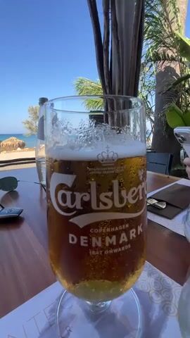 A close-up shot focuses on a tall glass of Carlsberg beer, condensation visible on the outside. The beer is a golden amber color with a frothy head. The Carlsberg logo, the word "Carlsberg," is prominently displayed in white lettering on the glass, along with "COPENHAGEN DENMARK 1847 ONWARDS." The glass sits on a wooden table. In the background, out of focus, a bottle of water is visible to the left, and palm fronds and what appears to be a building structure are to the right. Beyond that, a bright blue sky and a glimpse of a sandy beach with a straw umbrella and the ocean can be seen, suggesting an outdoor setting, possibly a beachside cafe or restaurant. A person's hand holding a smartphone is partially visible on the right side of the frame. The camera angle is low, looking up at the glass of beer.