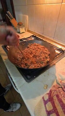 A person is seen in a kitchen, stirring a large quantity of small, brown, cooked pieces of meat or fat (possibly chicharrones) in a wide, dark pan resting on a gas stove. The pan occupies the central foreground of the frame.

The individual, whose arms and hands are visible, uses a long metal utensil to stir the contents of the pan. Another hand, partially visible on the left, holds a green-handled spatula. The activity suggests the process of cooking or rendering meat.

The setting is a kitchen with a light-colored tiled backsplash and a white countertop. On the left side of the counter, a white plate holding some food items (partially obscured) and a clear glass jar with a lid are visible. On the right, a colorful patterned towel is draped over the edge of the counter. The person's lower legs and white sneakers are visible at the bottom of the frame, indicating they are standing in front of the stove. The lighting suggests an indoor daytime setting or a well-lit interior. No visible text is present in the video.