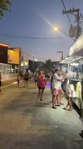 The video depicts a street scene in a tropical or coastal area at dusk or early evening. The sky is a blend of orange and blue. Several people are walking down the street, some in swimwear, indicating proximity to a beach or water. One man carries a child on his shoulders. 

A food truck or vendor stand is positioned on the right side of the street, with people gathered around it, presumably ordering or waiting for food. Buildings line the left side of the street, one with visible signage. A bright street light illuminates the scene, casting shadows. Power lines are visible overhead. The overall atmosphere is casual and relaxed, suggesting a vacation or leisure setting.