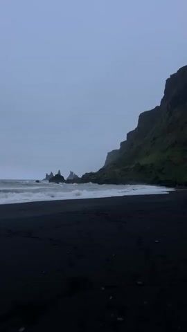 The video shows a black sand beach on a cloudy day.  To the left are several jagged rock formations in the ocean.  To the right is a cliff covered in green vegetation. Waves are breaking on the shore.  Footprints are visible in the black sand. The sky is overcast and gray.