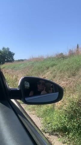A first-person perspective video captures a person driving a car on a sunny day. The camera is pointed at the side-view mirror, which reflects the driver taking a selfie with their phone. The driver is wearing sunglasses and a dark top. The background outside the car shows a grassy embankment and a clear blue sky. The car appears to be on a road with some gravel on the side. The video is short and seems to be a quick snapshot of a road trip.