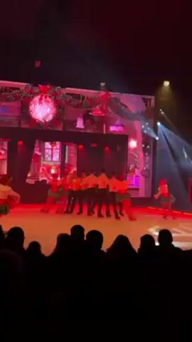 A group of ice skaters are performing on a stage in front of a live audience. The skaters, numbering about a dozen, wear matching outfits of orange shirts, black pants, and green ruffled skirts. They are arranged in a line, holding hands and skating in unison. Behind them, a large screen displays a holiday-themed image, featuring a decorated building with a festive wreath and lights. The scene is lit with red and white spotlights, enhancing the festive atmosphere. The audience is visible in the foreground as dark silhouettes, watching the performance. The stage is set up like a holiday street scene.