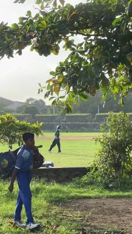 The video opens with a close-up of a young boy in a blue cricket uniform, carrying a cricket bag over his left shoulder. He is standing in a grassy area with dirt paths, looking towards his right. In the background, a cricket field is visible, with a few players in cricket attire practicing. One player is in the middle of the field, and another is further in the distance. The sky appears overcast. The scene is framed by lush green foliage from trees in the foreground and middle ground. The lighting suggests it might be early morning or late afternoon.