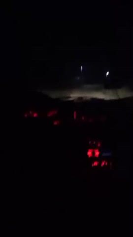 The video is a first-person perspective from inside a car at night. The dashboard lights are illuminated in red. The headlights of the car are on and are illuminating a dirt or gravel road ahead. There are some distant lights visible in the darkness, possibly from buildings or streetlights. The car appears to be moving forward.