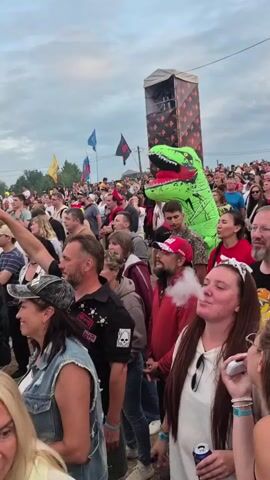 The video shows a large crowd of people at an outdoor event. A giant inflatable green dinosaur costume is visible in the middle of the crowd, with a stage or platform behind it. Many people are looking in the same direction, presumably at a performance or attraction. Some individuals in the foreground are smoking. The sky is cloudy.