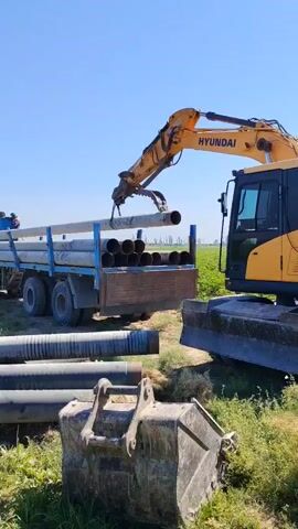 A construction scene is shown on a clear day with a bright blue sky. A yellow Hyundai excavator is positioned to the right of the frame, its arm extended to lift a long, gray pipe from a flatbed truck. The truck, also blue, is loaded with similar pipes. In the foreground, a large excavator bucket sits on the ground amidst green grass and some dirt. Several other pipes are also on the ground in front of the truck. A person can be seen on the back of the truck, assisting with the pipe loading. The environment appears to be rural or agricultural, with a field of crops visible in the background.