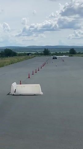 A dark blue hatchback car is seen driving on an asphalt track, maneuvering between orange traffic cones. The car appears to be in motion, with its wheels turned as it navigates the course. In the foreground, a white plastic barrier is positioned on the track. The background shows a landscape with rolling hills, vegetation, and a cloudy sky. There are also some structures visible in the distance, including a small building and what appear to be additional traffic cones. The overall setting suggests a driving course or test track.