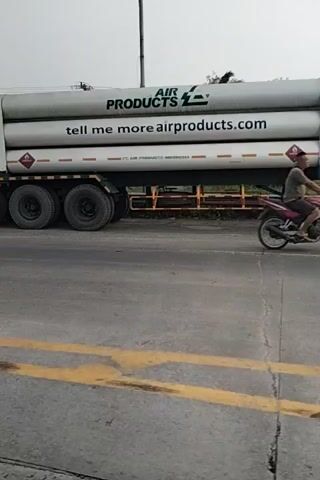The video is a first-person perspective of a truck carrying large cylindrical tanks. The truck is moving on a paved road with a yellow line. The tanks are white and labeled with text. One tank reads "AIR PRODUCTS". Another tank reads "tell me more airpro". A smaller tank reads "HYDROGEN COMPRESSED". Another smaller tank reads "DILARANG MEROKOK". The truck has multiple wheels visible. The environment appears to be outdoors with vegetation, trees, and a cloudy sky. In the background, there are some structures that resemble small houses or shelters and a power pole with wires. The camera is shaky, indicating movement.