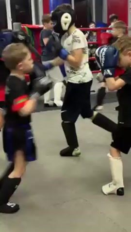 In a gym setting, several young children, dressed in protective gear including headgear, gloves, and shin guards, are engaged in a boxing sparring session. They are paired off and throwing punches at each other, moving around on a gray padded floor. A boxing ring is visible in the background with red, white, and blue ropes, along with other training equipment such as punching bags. The walls are a combination of red and white. The atmosphere appears to be focused on training and practice.