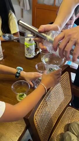 The video shows a group of people, including children and adults, gathered around a wooden table. In the foreground, a person's arm is visible, holding a clear plastic cup with a green substance and ice cubes. Another person's arm is seen pouring a clear liquid from a metal dispenser into a similar cup. There are several other clear plastic cups on the table, some containing liquid and ice, and some appearing empty. A water bottle with a panda design is also on the table, as is a spoon. In the background, a young child in a blue shirt is leaning over the table, looking into a cup. The overall environment appears to be a casual indoor setting, possibly a cafe or home. The activity seems to involve preparing or consuming drinks.