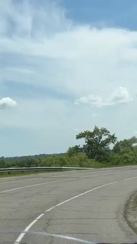 The video shows a view from inside a vehicle traveling on a paved road. The road has white lane markings. On the left side of the road is a metal guardrail and a grassy embankment covered with green bushes and trees. The sky is blue with scattered white clouds. On the right side of the road, there is a grassy slope with trees. The road curves to the right. In the distance, there are more trees and some buildings. The video appears to be filmed on a sunny day.