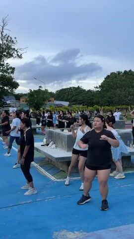In an outdoor setting with a blue painted ground and scattered benches, a group of people are engaged in a Zumba class. The time of day appears to be late afternoon or early evening, with the sky displaying a mix of clouds and hints of twilight. The weather seems mild, as participants are dressed in athletic wear such as tank tops, t-shirts, shorts, and leggings.

The subjects are primarily women, though a few men are also visible in the background. They are arranged in rows, facing a common direction, suggesting an instructor is leading the session, though the instructor is not clearly visible in this frame. Their postures and movements indicate active participation in a choreographed dance fitness routine, with some individuals focused and others seemingly enjoying the activity. The overall mood appears energetic and positive.

The background features lush green trees and some low-rise buildings, hinting at a park or an open community space within a populated area. The presence of streetlights further confirms an urban or semi-urban environment. No visible text is present in the provided frame.