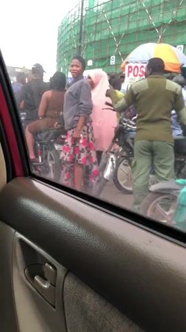 The video captures a street scene with multiple motorcycles and a yellow bus. In the foreground, a person is seen through a car window, looking out at the street. A woman in a grey sweatshirt and a patterned skirt stands on the street, facing the camera. Behind her, a person is seated on a motorcycle wearing a brown outfit. Several other motorcycles with riders are visible, along with a larger white and teal bus. The background shows a construction site with green scaffolding. The scene appears to be a busy street, possibly in a city, with people and vehicles engaged in various activities.