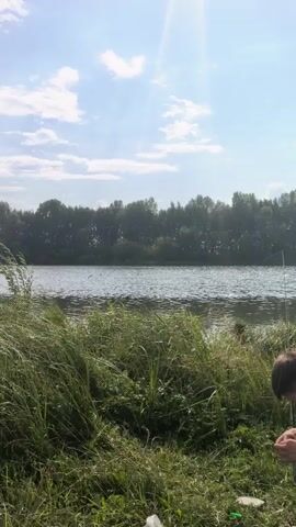 A person is fishing by a lake. The sun is shining brightly in the sky, and there are clouds present. The lake is calm, with trees lining the opposite shore. The person is kneeling on the bank of the lake, tending to their fishing gear. Tall green grass and reeds are visible in the foreground.
