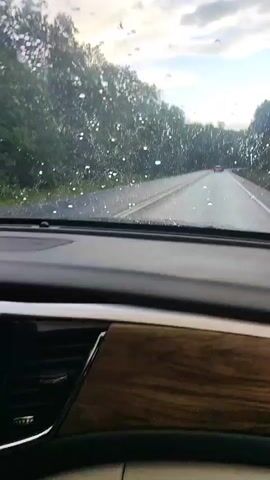 This video shows the interior of a car, focusing on the dashboard and infotainment system. The windshield is visible in the background, with raindrops on it, suggesting it is raining. The infotainment screen displays multiple widgets, including a clock, a navigation interface showing a car icon on a road, and a music player with a musical note icon. Below the infotainment screen is the climate control panel with various buttons and dials. A hand is seen reaching for something near the gear shift. The overall impression is that of a person inside a vehicle during inclement weather, possibly looking at their car's media or navigation system.
