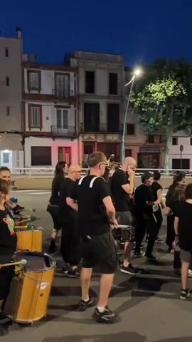 A group of people in black clothing are playing drums and percussion instruments on a street at dusk. The buildings in the background appear to be old, with balconies and shutters. A streetlamp illuminates the scene. The people are of various ages and genders, and they are all facing in the same direction, as if marching or performing. One person in the foreground is playing a large drum, while others are playing smaller drums and what appear to be snare drums and cymbals. The overall atmosphere is one of a lively outdoor performance or parade.