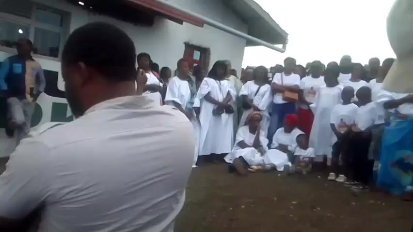Here is a description of the video:

The video appears to show a gathering of people outside a building. The people are of diverse ages, with many adults and some children. A number of the adults are wearing long white robes, while others are wearing t-shirts or casual attire. A few children are wearing t-shirts that have an image on them. Some women are seated on the ground. A man in a striped shirt is standing in the foreground, facing the group, while another man with a baseball cap is partially visible on the right side of the frame. The building behind the group is white, with at least one window and a red trim along the roofline. The ground is bare earth. The people appear to be gathered for an event or ceremony, but the exact nature of it is unclear from the video alone.