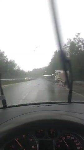 The video shows a first-person perspective from inside a car driving on a wet road during a rainstorm. The windshield wipers are intermittently activated. The road is bordered by trees on both sides, and there are other vehicles visible in the distance, including a truck. The sky is overcast and gray, indicating heavy rain or bad weather. The road surface is slick and shows reflections of the sky and vehicles. There are also some debris on the road, possibly from the storm.