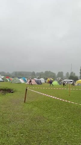Three people in athletic wear are running across a grassy field. The sky is overcast and appears to be raining. In the background, there are numerous tents set up, suggesting a campsite or event. There are also trees and some buildings visible in the distance. Red and white striped tape is stretched across the foreground, indicating a boundary or path. The ground is wet and appears muddy in places.