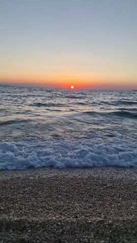 The video shows a sunset over a beach. The sun is a bright orange ball low on the horizon. The sky is a gradient of orange, pink, and blue. The ocean has moderate waves with white foam. The beach is covered in small, gray and white pebbles. There are no people or objects visible in the scene.