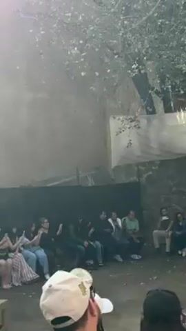 The video shows an outdoor performance with an audience seated in front of a stone wall. A man dressed in a green robe and a crown is performing for the audience. He walks back and forth, gesturing with his arms. A white awning provides some shade. Some of the audience members are clapping. Trees are visible above the wall, casting shadows on the scene. A person in the foreground appears to be filming the performance.