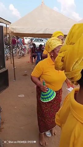 The video shows a group of middle-aged African women dancing at an outdoor event. All are wearing matching yellow tops and head wraps, and printed reddish-brown skirts. One woman in the foreground is holding a fan and a white cloth. The women are standing on bare dirt. In the background, there is a white tent, chairs, vehicles, and other people sitting and standing around. The video appears to be shot from the perspective of someone participating in the dance. The sound of music and singing can be heard.