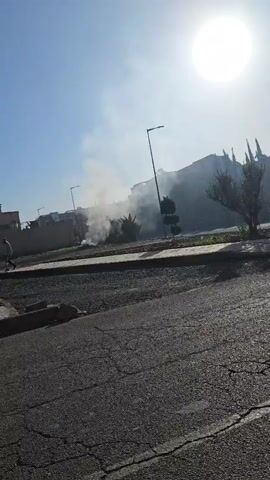 The video shows a street scene on a sunny day. Smoke is visible in the distance, near trees and buildings. The camera pans across the street, showing a sidewalk, light poles, and a small utility box. The street is cracked and shows signs of wear. There are no people visible in the video. The sky is blue with a bright sun.