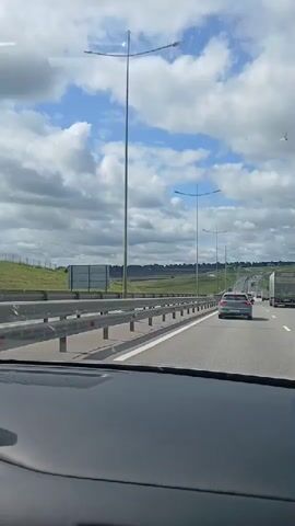 A view from inside a vehicle traveling on a highway under a partly cloudy sky. The road has multiple lanes with other vehicles, including cars and trucks. A metal guardrail separates the highway from a grassy area and fields beyond. The sky is filled with white, puffy clouds against a blue background.