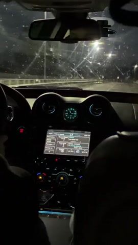 The video captures an interior view of a car at night, filmed from the passenger seat looking forward. A person's left hand is visible on a dark steering wheel, indicating a driver. The dashboard is illuminated, featuring a central analog clock flanked by two circular air vents, and a large touchscreen display below them. Further below are physical buttons and knobs. A rear-view mirror and a dashcam are visible attached to the windshield. The windshield is covered with numerous small water droplets or smudges. Outside, streetlights illuminate a road ahead, suggesting the car is driving on an elevated highway or bridge at night. Briefly, a right hand enters the frame from the bottom left, pointing towards the dashboard.