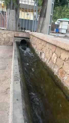 The video shows a long, narrow concrete channel filled with murky green water flowing from left to right. The channel is bordered by a stone wall on the right and a concrete ledge on the left. A pipe enters the channel from the left, discharging the water. In the background, there are some trees, a metal fence, and what appears to be a small building or structure. The water is moving, creating ripples and small splashes. The overall scene suggests an outdoor drainage or irrigation system.