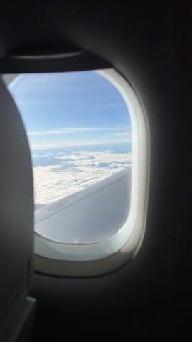 The video is an aerial view from inside an airplane, looking out the window. The perspective is from a passenger seat, with the dark interior of the plane visible on the left and right edges of the frame. The primary focus is the vast expanse of white clouds spread out below, resembling a soft, undulating landscape. Above the clouds, the sky is a clear, bright blue with a few faint wispy clouds. The wing of the airplane is visible in the lower right portion of the frame, extending towards the horizon. The sunlight illuminates the scene, creating a bright and serene atmosphere. The view suggests the airplane is flying at a high altitude.