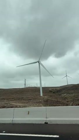 The video shows a close-up view of a wind turbine against a cloudy sky. The turbine is white with three blades, and the blades are in motion. In the background, there are other wind turbines, as well as a power tower and some vegetation. The foreground is dominated by a white concrete barrier and a dark road surface, suggesting the footage was taken from a vehicle. The overall impression is one of a remote, industrial landscape with a focus on renewable energy.