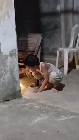 A young boy is crouching on a concrete floor, looking at something on the ground near a wall. He is wearing a light-colored tank top and patterned shorts. To the left, there is a wall with some discoloration and a wooden structure. To the right, there is a white plastic chair. The lighting is dim, with a bright light source illuminating the area where the boy is looking. The boy appears to be interacting with an object on the ground.