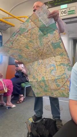 A man wearing a headset microphone and an olive green vest over a black t-shirt and blue jeans stands in a subway car, holding up a large, unfolded map of Moscow with both hands. He lowers the map, revealing a mustache and a slight smile. He then folds the top part of the map downwards, showing a map of the Moscow region on the back. Other passengers are visible in the background, seated.