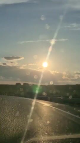 The video shows a view from inside a vehicle, looking out at a sunset. The sun is bright and partially obscured by clouds. A road is visible in the foreground, and a field or landscape stretches out to the horizon. The windshield has water droplets and streaks, which create light flares and bokeh effects, obscuring some of the details of the scene. The lighting suggests it is late afternoon or early evening.