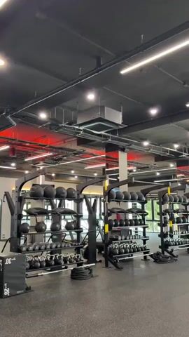 This video pans across a modern, well-equipped gym. The gym features black rubber flooring and a dark ceiling with exposed pipes and modern LED lighting. Several racks of dumbbells, kettlebells, and medicine balls are visible. In the background, large windows offer a view of greenery. There are also squat racks with weight plates and barbells. The overall impression is a clean, spacious, and functional fitness facility. There are no people visible in the frame.
