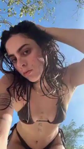 A young woman with dark, wet hair and a dark brown bikini stands outside, likely at the beach, with a bright blue sky and tree branches visible in the background. The camera angle is low, looking up at her. She appears to be recording herself, adjusting her hair and briefly sticking out her tongue toward the camera. She has tattoos on her chest and arm. The atmosphere seems sunny and relaxed.