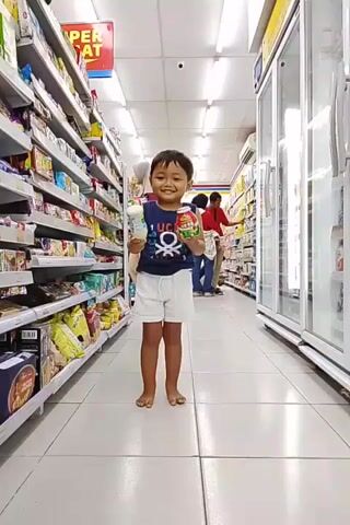 A young boy is walking down an aisle in a brightly lit convenience store. He is wearing a dark blue sleeveless shirt with a white graphic and white shorts. He is barefoot. In his right hand, he holds a small, round, red and white container, possibly a snack or drink. In his left hand, he holds a white bottle, also possibly a drink or condiment. The boy is smiling and looking towards the camera. To his left, shelves are stocked with various products, including snacks and packaged goods. To his right, there are glass-fronted refrigerators. Other shoppers are visible in the background, further down the aisle. The floor is tiled in a white and light gray checkerboard pattern.