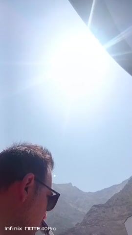 A man wearing sunglasses and an orange vest is in the foreground, with mountains in the background. The sun is shining brightly, creating lens flare. The man turns his head towards the camera.