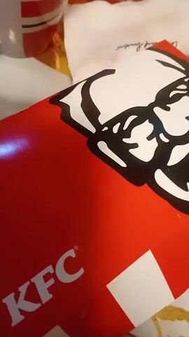 A hand with long, red painted nails is shown opening a red and white KFC box. Inside the box are several pieces of fried chicken with a golden-brown crispy breading, and a small red ketchup packet. The box is sitting on a table, and a paper towel and a white mug are visible in the background.