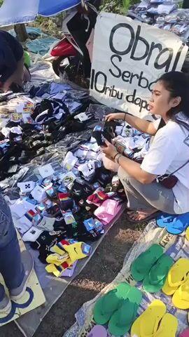 This video shows an outdoor market scene, likely a roadside stall. Several people are present, including women and possibly children. They are gathered around a display of goods laid out on the ground, covered by tarps and cloths. The items appear to be primarily socks, with various colors, patterns, and brands visible. Some women are seen examining the socks, suggesting they are customers or vendors. A sign with writing in Indonesian, "Obrol Serba 10 ribu dapet" (loosely translated as "Sale Everything 10 thousand get"), indicates that the items are being sold at a promotional price. Flip-flops in green and yellow are also visible in the foreground. In the background, motorcycles are parked, and a white umbrella provides shade. The environment appears to be open, with hints of greenery and a bright sky, suggesting it is daytime. The overall atmosphere suggests a busy, informal marketplace.